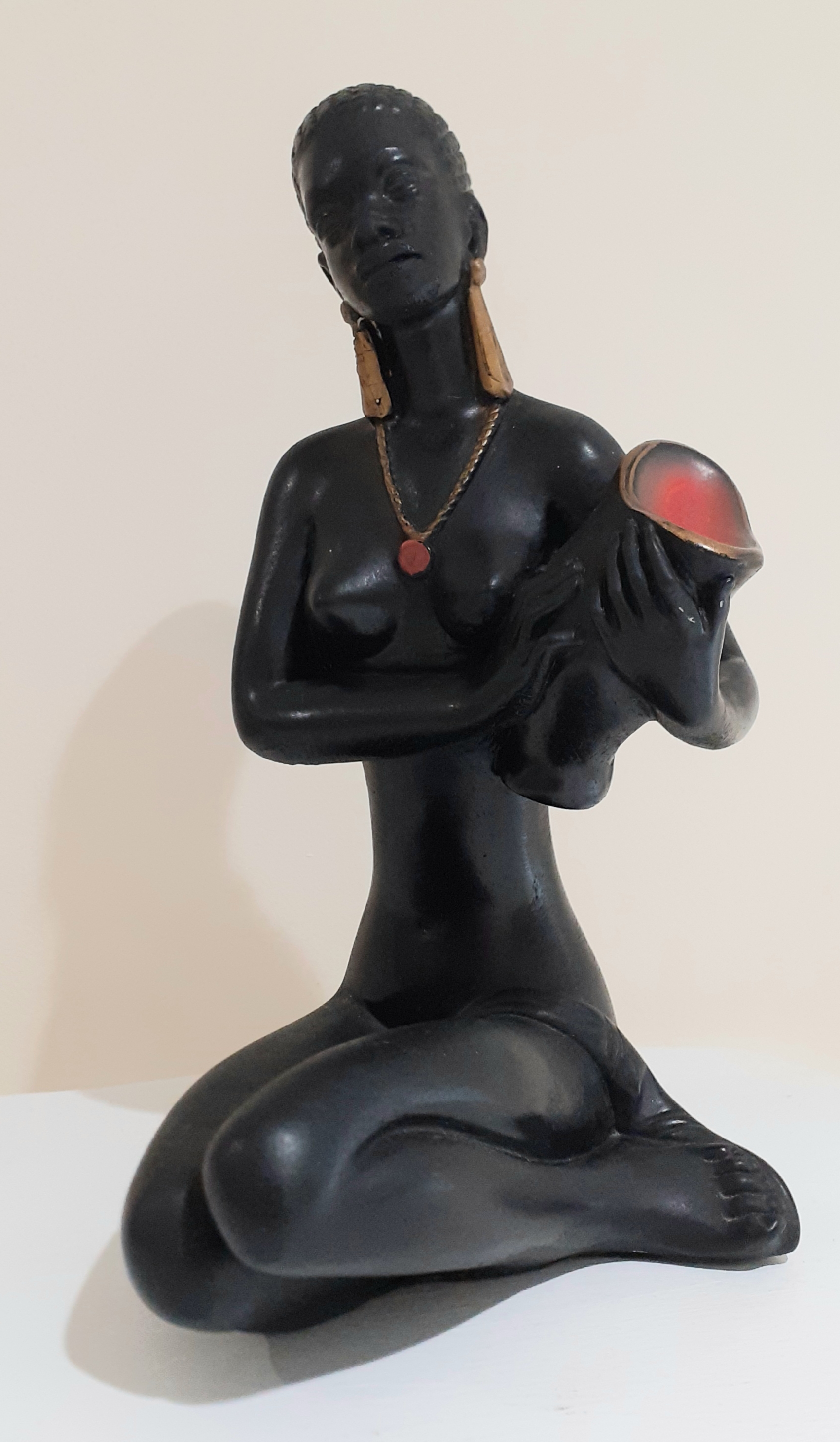 Kneeling lady figurine made of chalkware holding a black vase with red top close to the left hand side of her chest